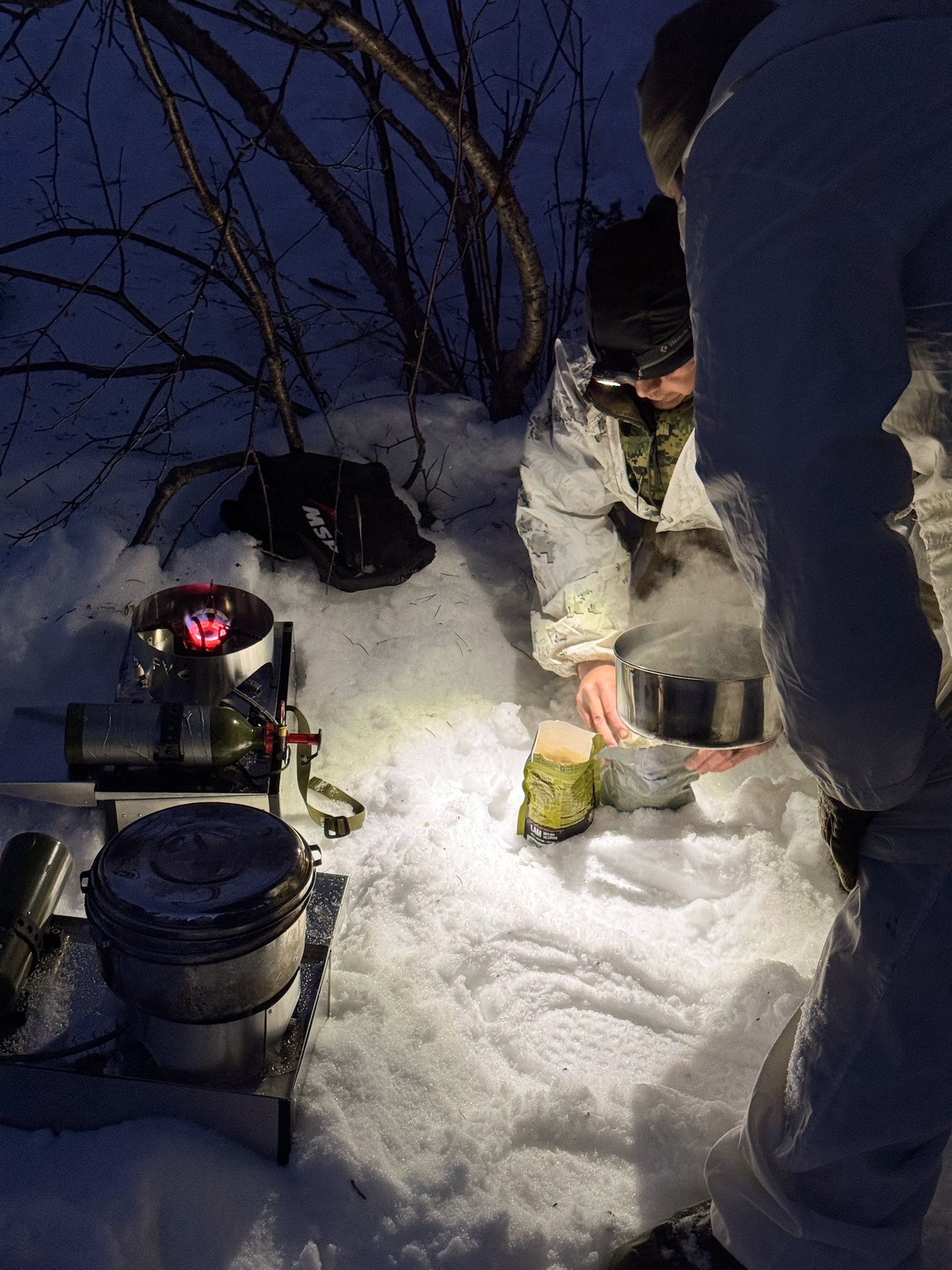 Participants cooking by headlamp after dark, with a lit Fyringsvakta stove and a pot on the snow.
