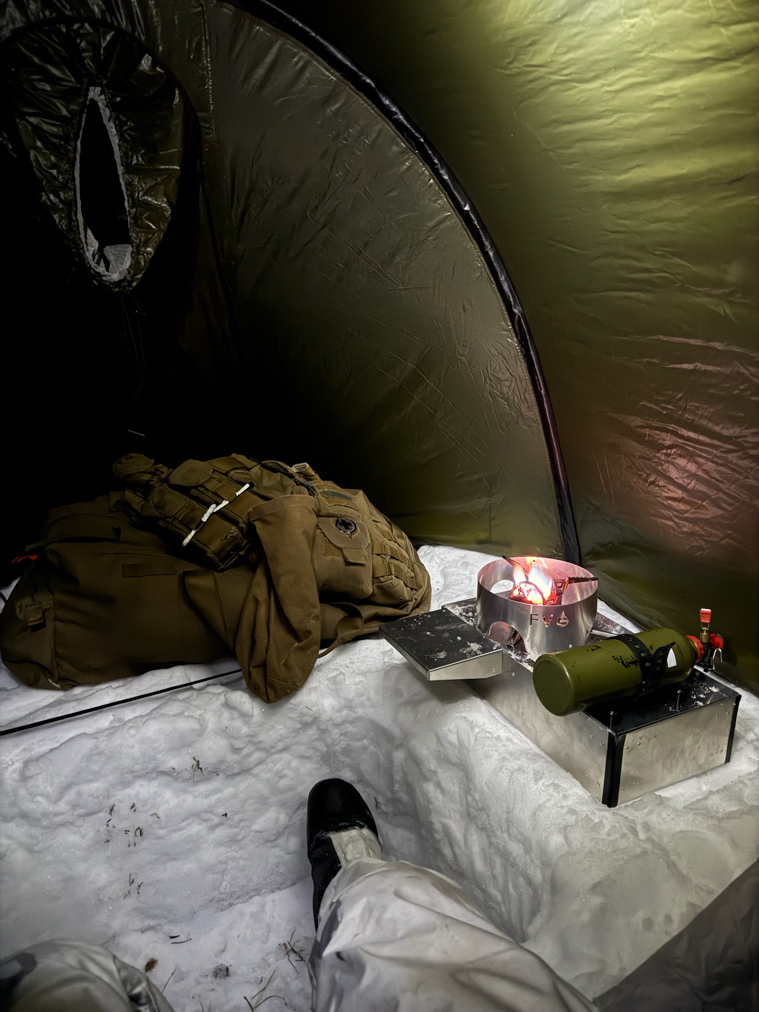 A lit Fyringsvakta stove on the snow floor of a green tent, next to a pack and load-carrying vest.