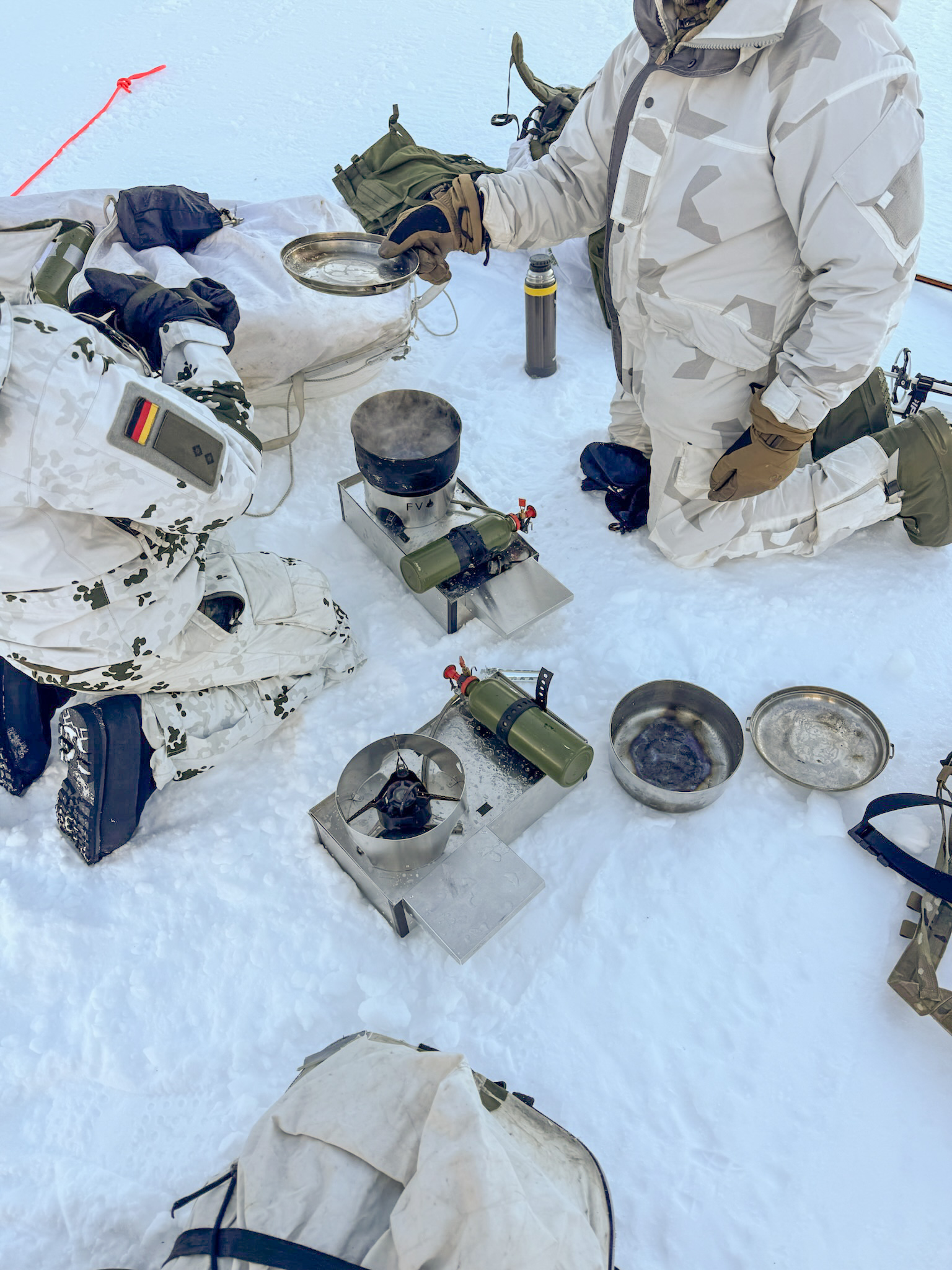 Overhead view of two Fyringsvakta stoves in use on the snow, with participants kneeling on either side.