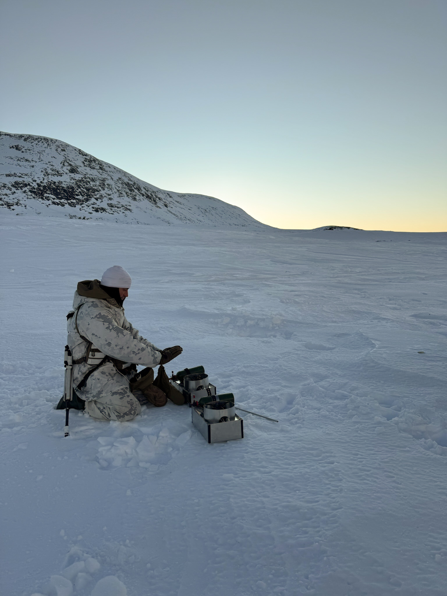 A participant kneeling beside two Fyringsvakta stoves on a snow-covered plateau under a pale sky.