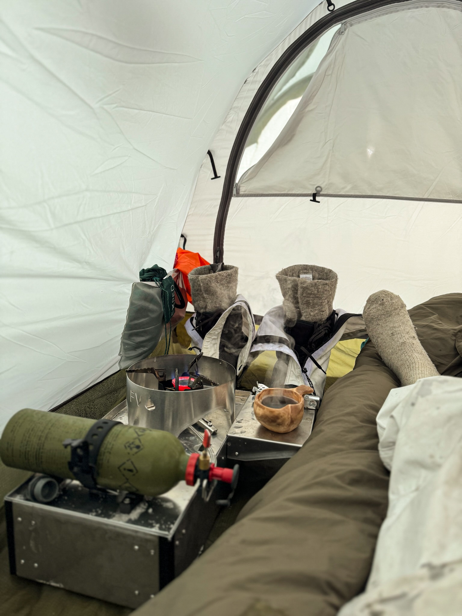 Inside a tent: a lit Fyringsvakta stove, a steaming wooden cup, boots and a sleeping bag.